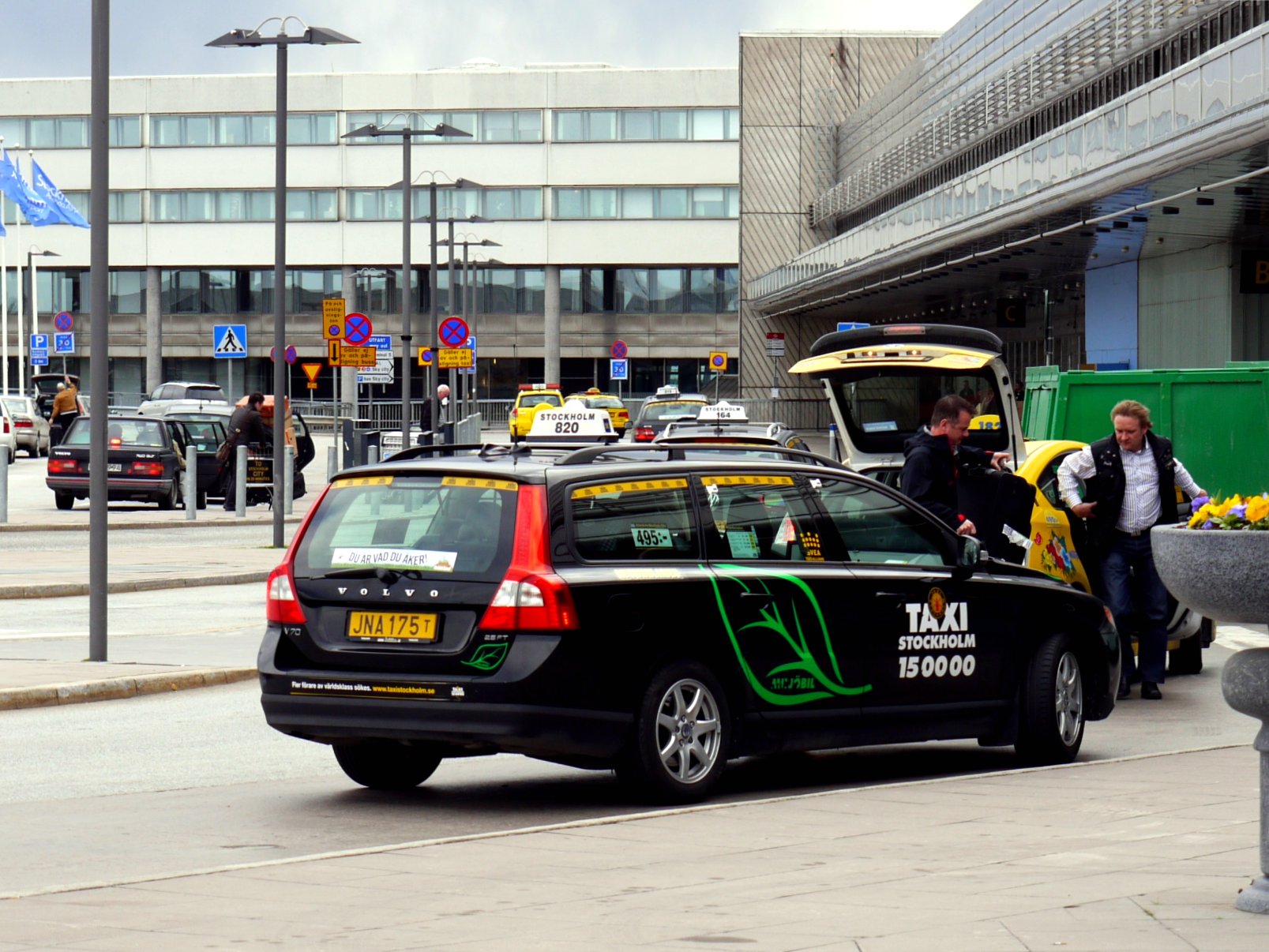 A black Taxi Stockholm estate car loading luggage at the taxi rank outside an Arlanda Airport terminal, with yellow taxis behind