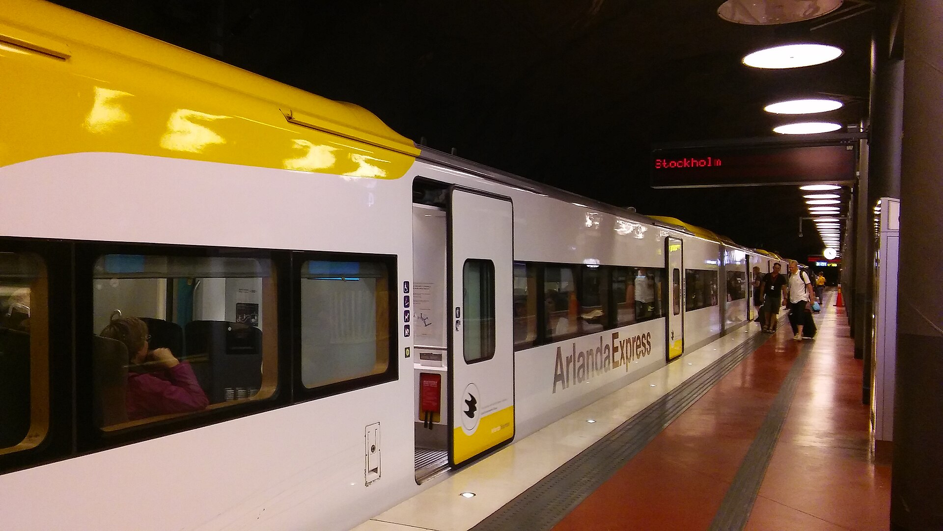 A white-and-yellow Arlanda Express train at an underground platform with its door open and passengers with suitcases walking along the platform