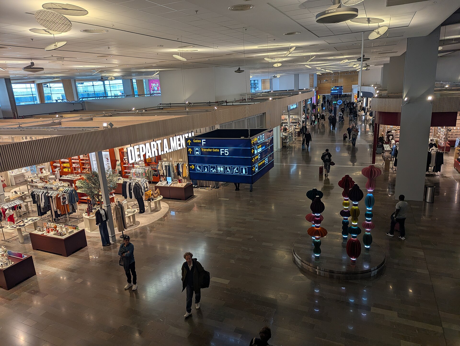The airside shopping area of Arlanda Terminal 5 seen from above, with shops, a departures board and travellers walking through