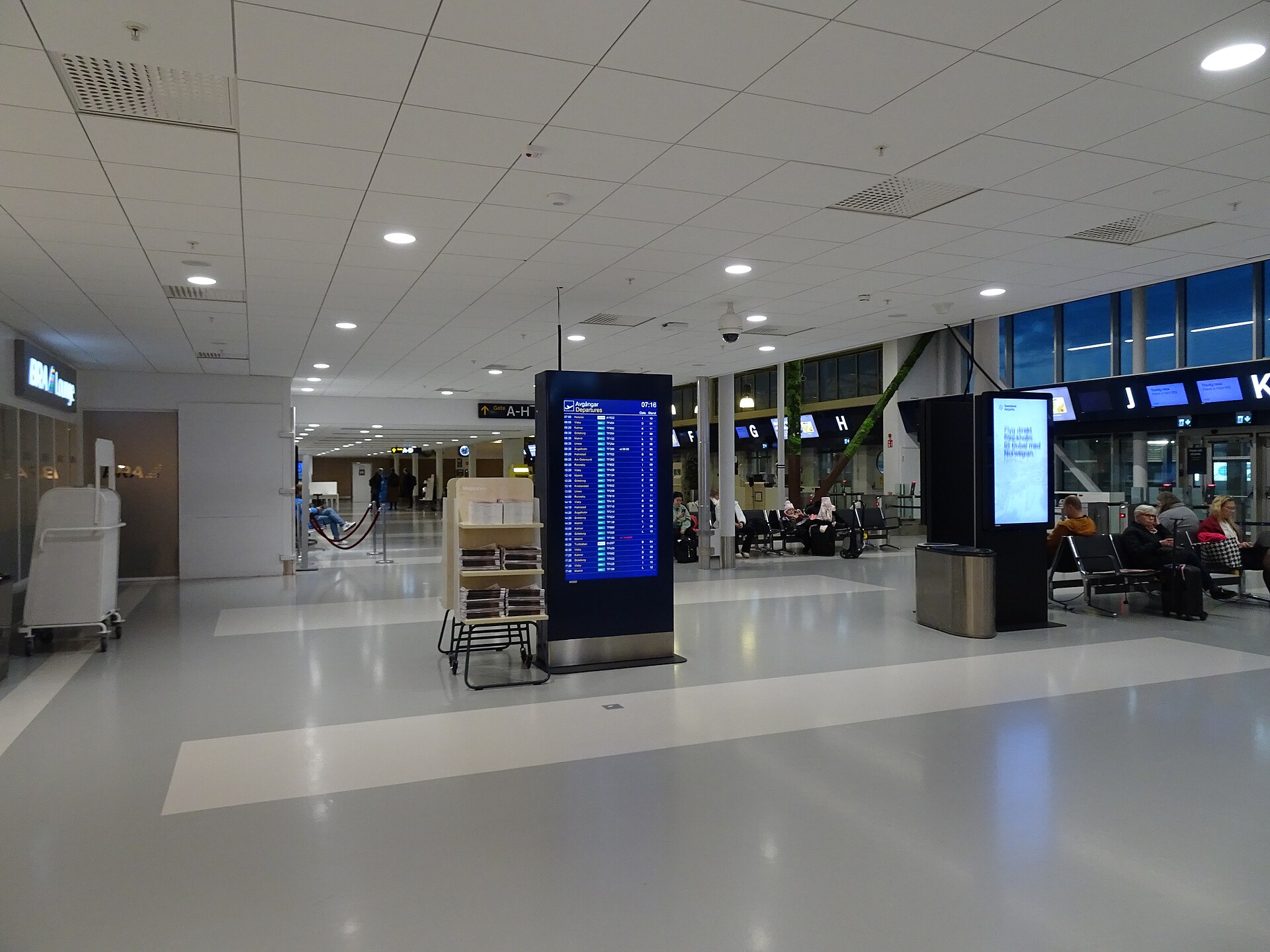 The quiet departure hall at Bromma Airport with a blue departures board, check-in desks marked with letters and a few passengers seated by the windows