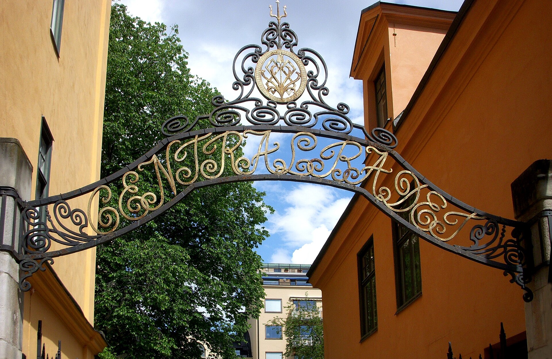 An ornate black and gold wrought-iron arch spelling Centralbadet over the entrance lane between ochre and orange buildings, with a leafy tree behind