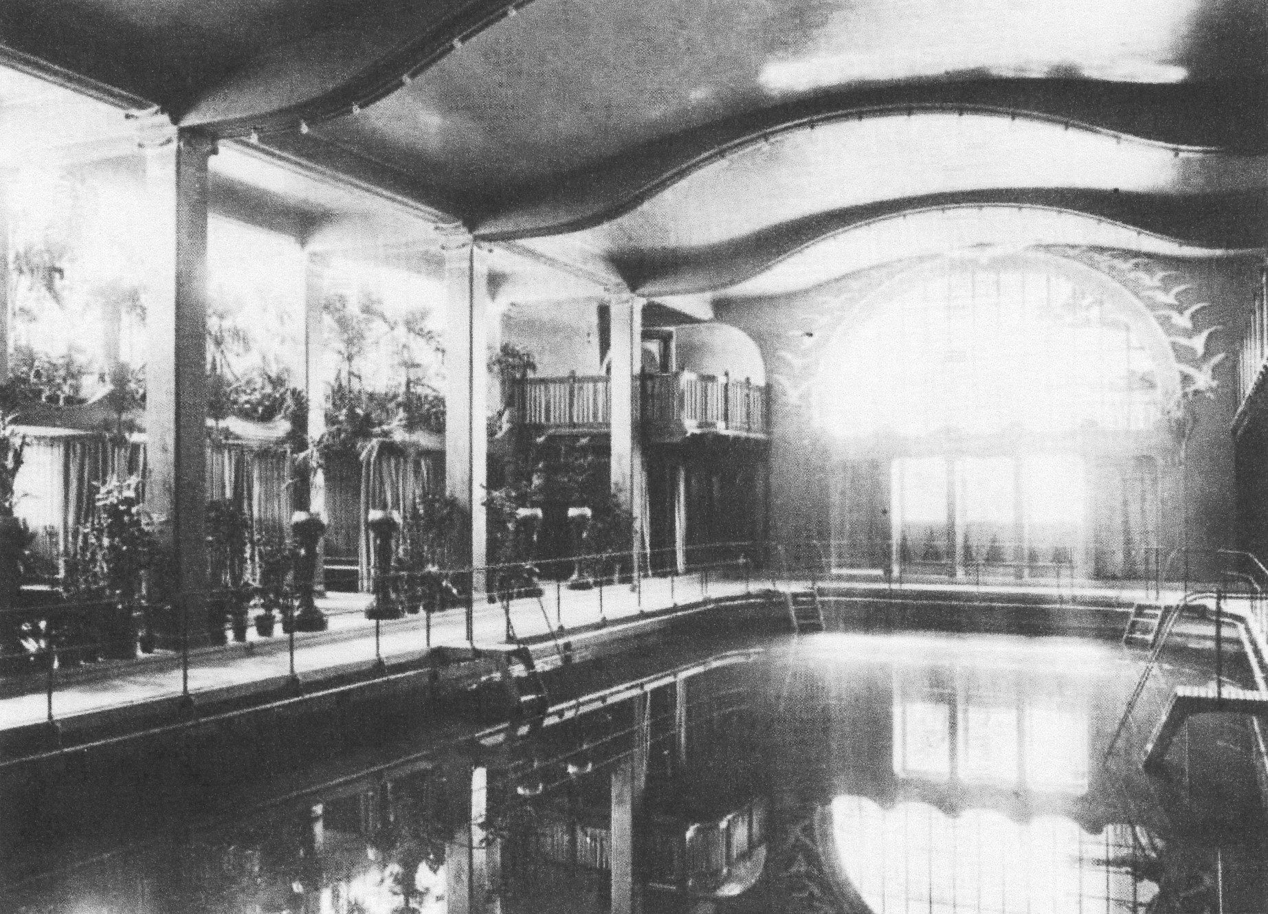 Black-and-white photograph of Centralbadet's pool hall in 1906, with a wavy Art Nouveau ceiling, potted palms, changing cubicles and a painted frieze of flying birds above a tall window