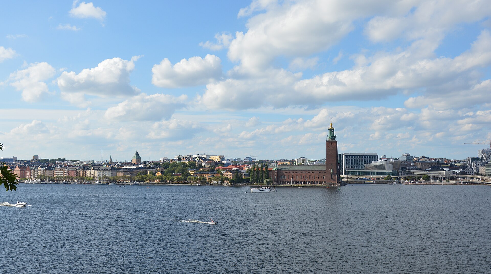 View across the wide water of Riddarfjärden to Stockholm City Hall's brick tower and the Kungsholmen shoreline under a blue sky with scattered cloud