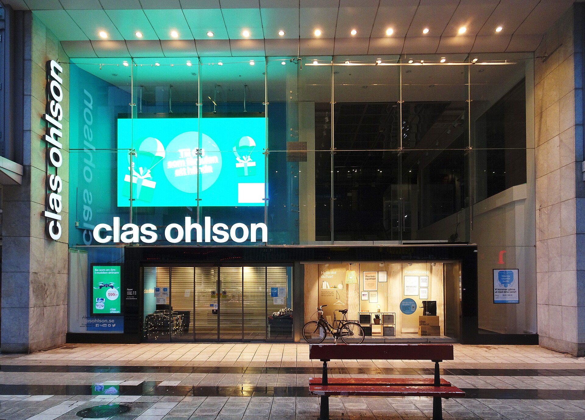 The illuminated glass frontage of a Clas Ohlson store on Drottninggatan in Stockholm at dusk, with a bench outside