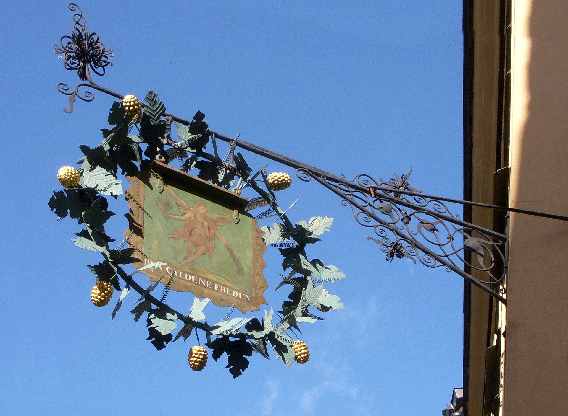 The wrought-iron hanging sign of Den Gyldene Freden in Gamla Stan, a painted panel framed by metal vine leaves and grapes against a blue sky