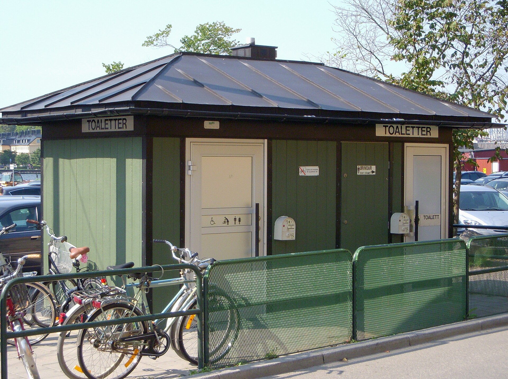 A small green wooden public toilet building with separate doors, bicycles parked outside and a green railing, on Djurgården