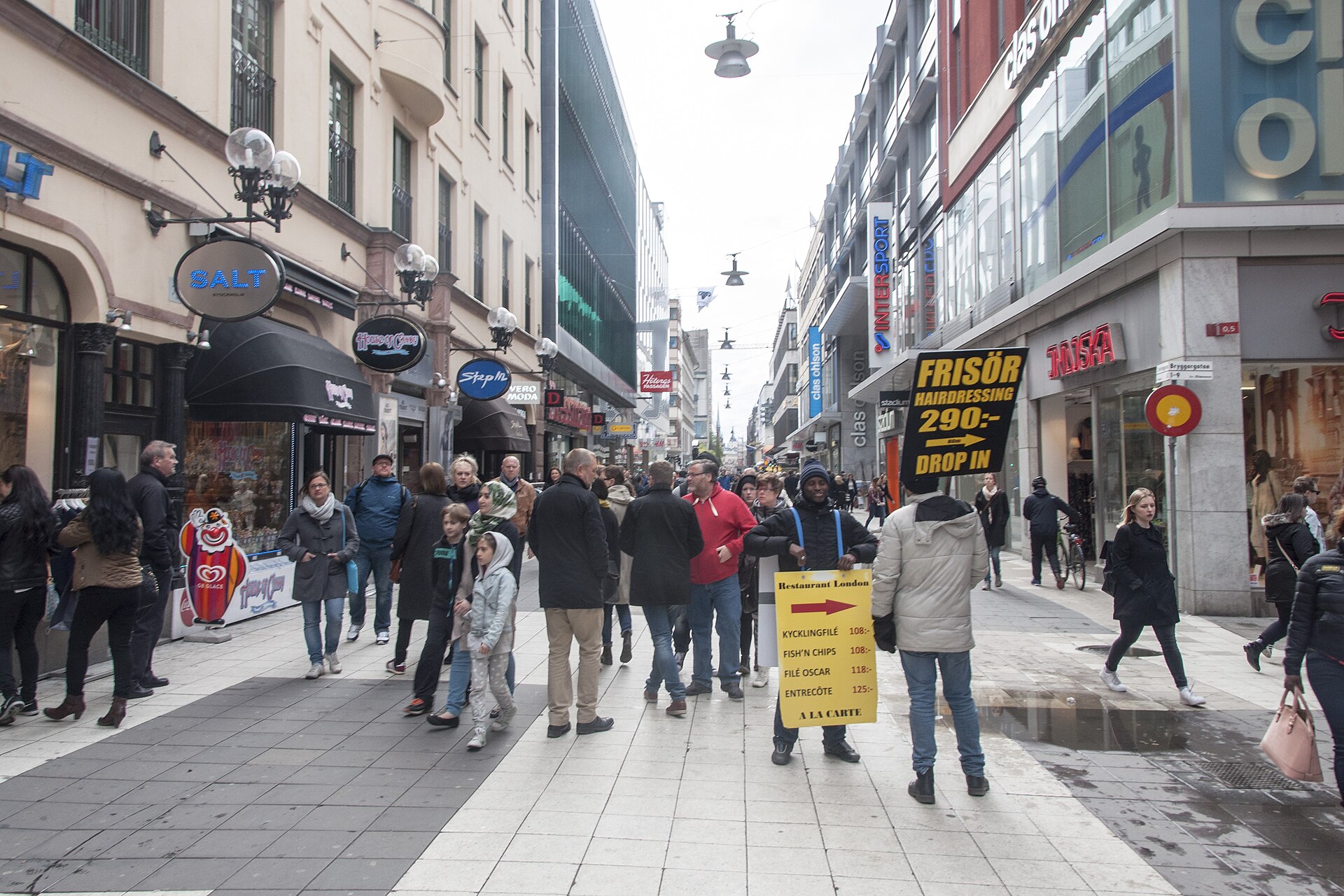 Crowds of shoppers on Drottninggatan in Stockholm, with shop signs and a man holding a hairdresser's advertising board