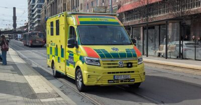 A yellow-and-green Swedish ambulance on a Stockholm street beside tram tracks, with shops and pedestrians behind