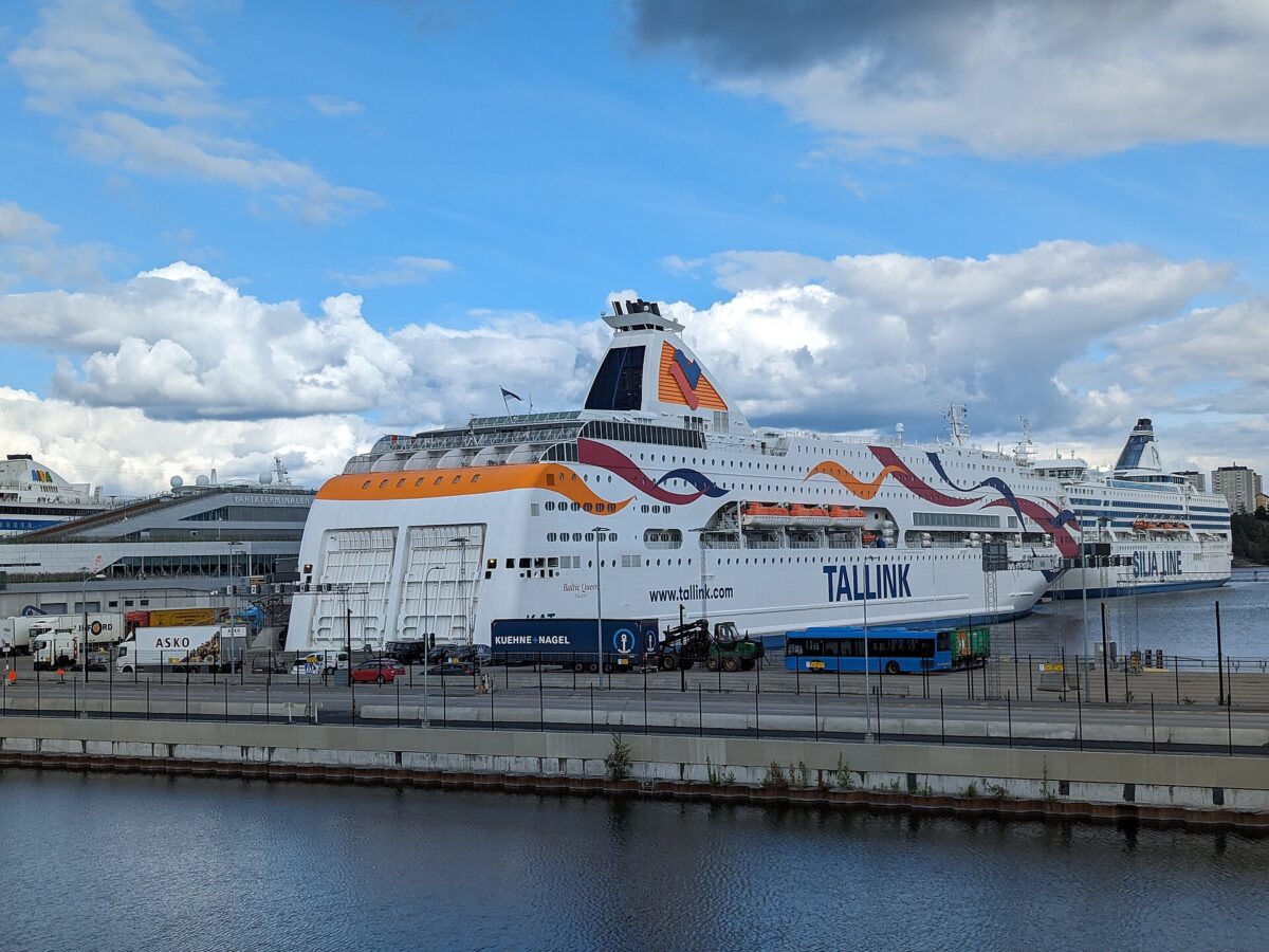 Two large white Baltic cruise ferries, Tallink's Baltic Queen and Silja Line's Silja Symphony, moored at the Värta ferry terminal in Stockholm under a blue sky with big clouds