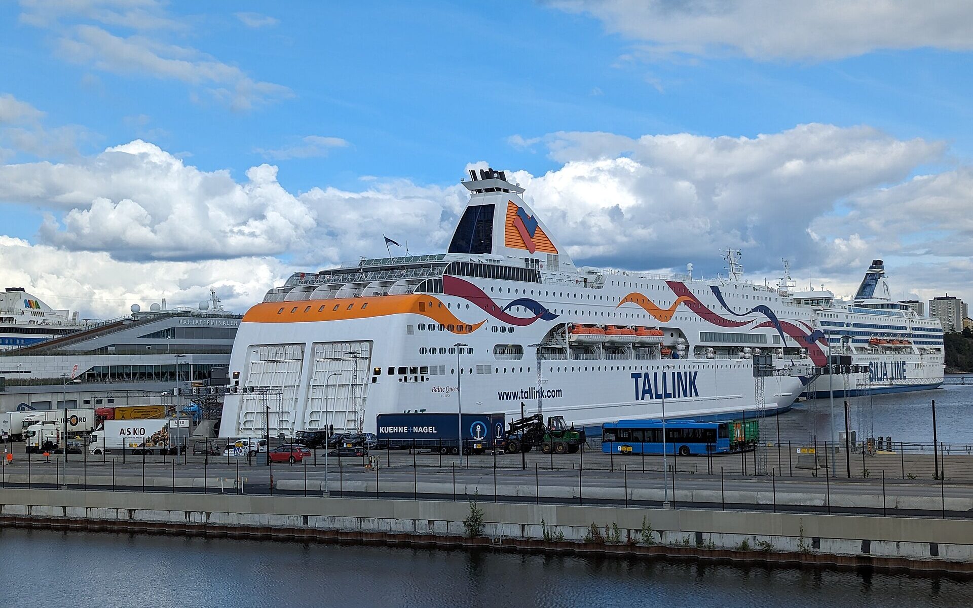 Two large white Baltic cruise ferries, Tallink's Baltic Queen and Silja Line's Silja Symphony, moored at the Värta ferry terminal in Stockholm under a blue sky with big clouds