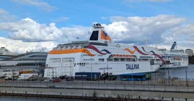 Two large white Baltic cruise ferries, Tallink's Baltic Queen and Silja Line's Silja Symphony, moored at the Värta ferry terminal in Stockholm under a blue sky with big clouds