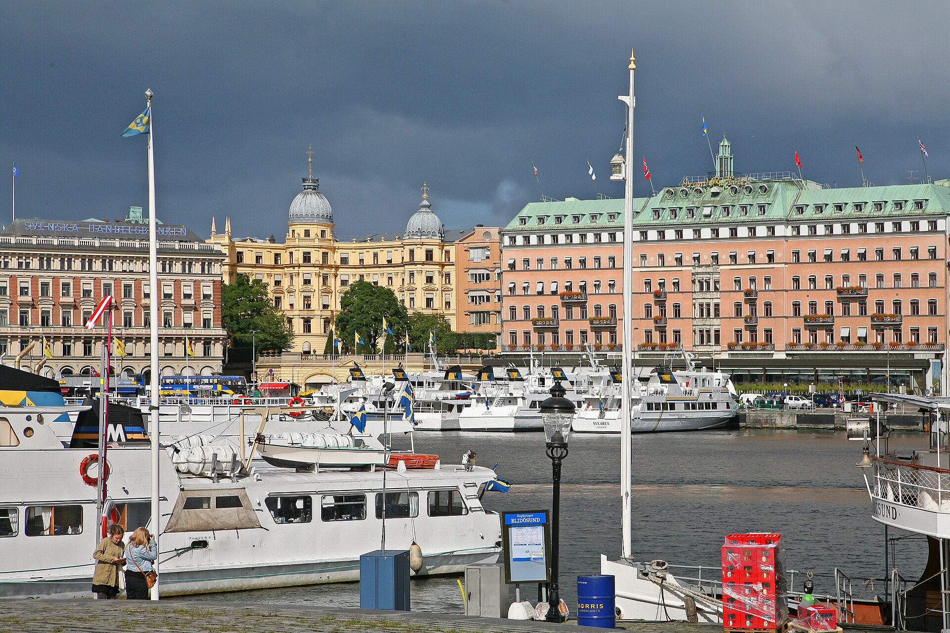 Grand Hôtel on the Blasieholmen waterfront in Stockholm, a long pale building with a green copper roof, with white passenger boats moored at the quay in front under a heavy grey sky