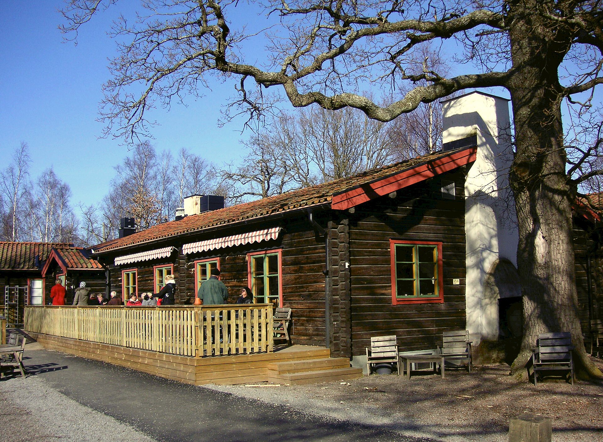 The dark-timbered lodge at Hellasgården with red window frames and a sunny wooden terrace where visitors gather, under a large bare oak tree
