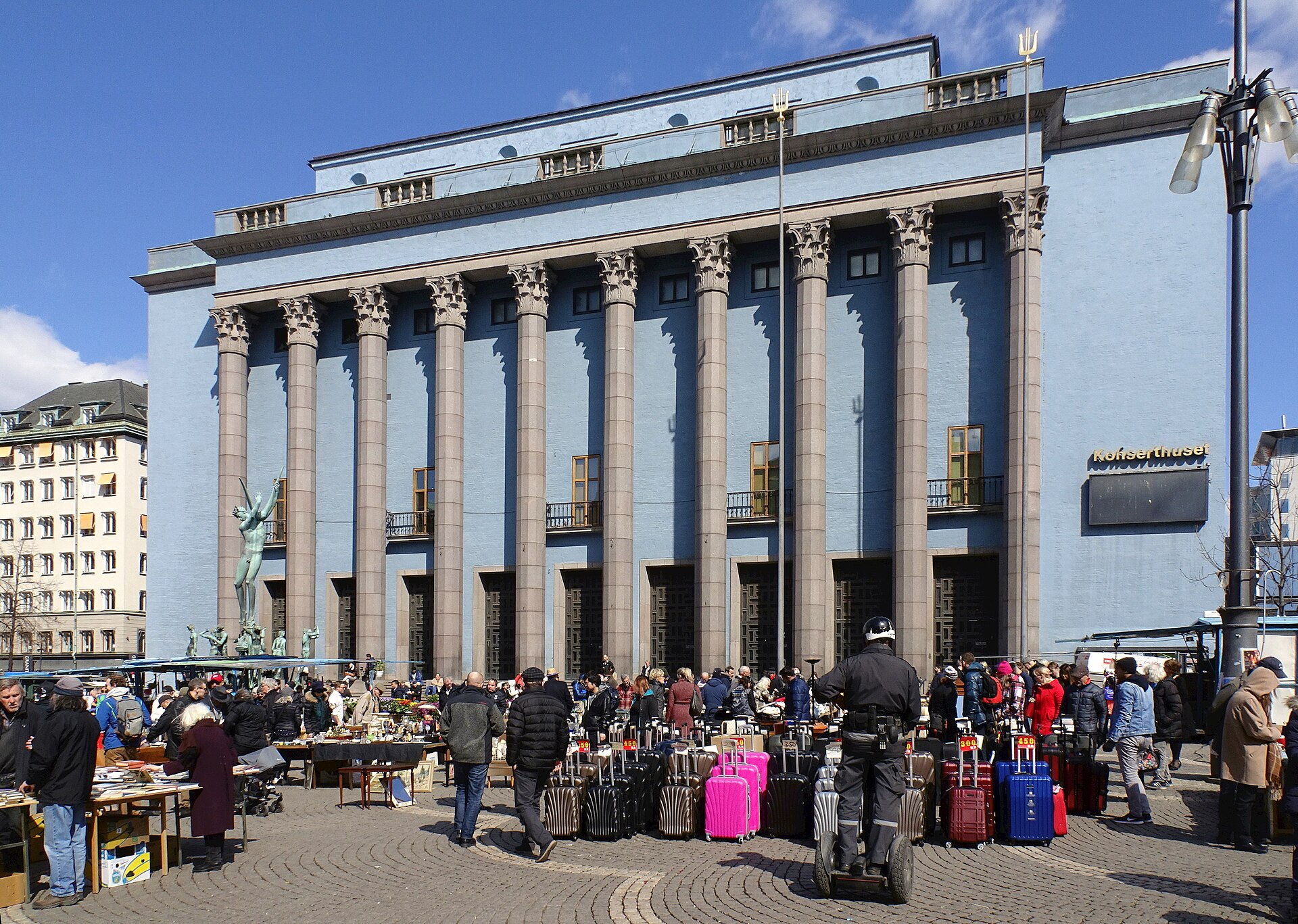 A crowd browsing flea market tables and a row of suitcases on Hötorget square in front of the blue columned Konserthuset