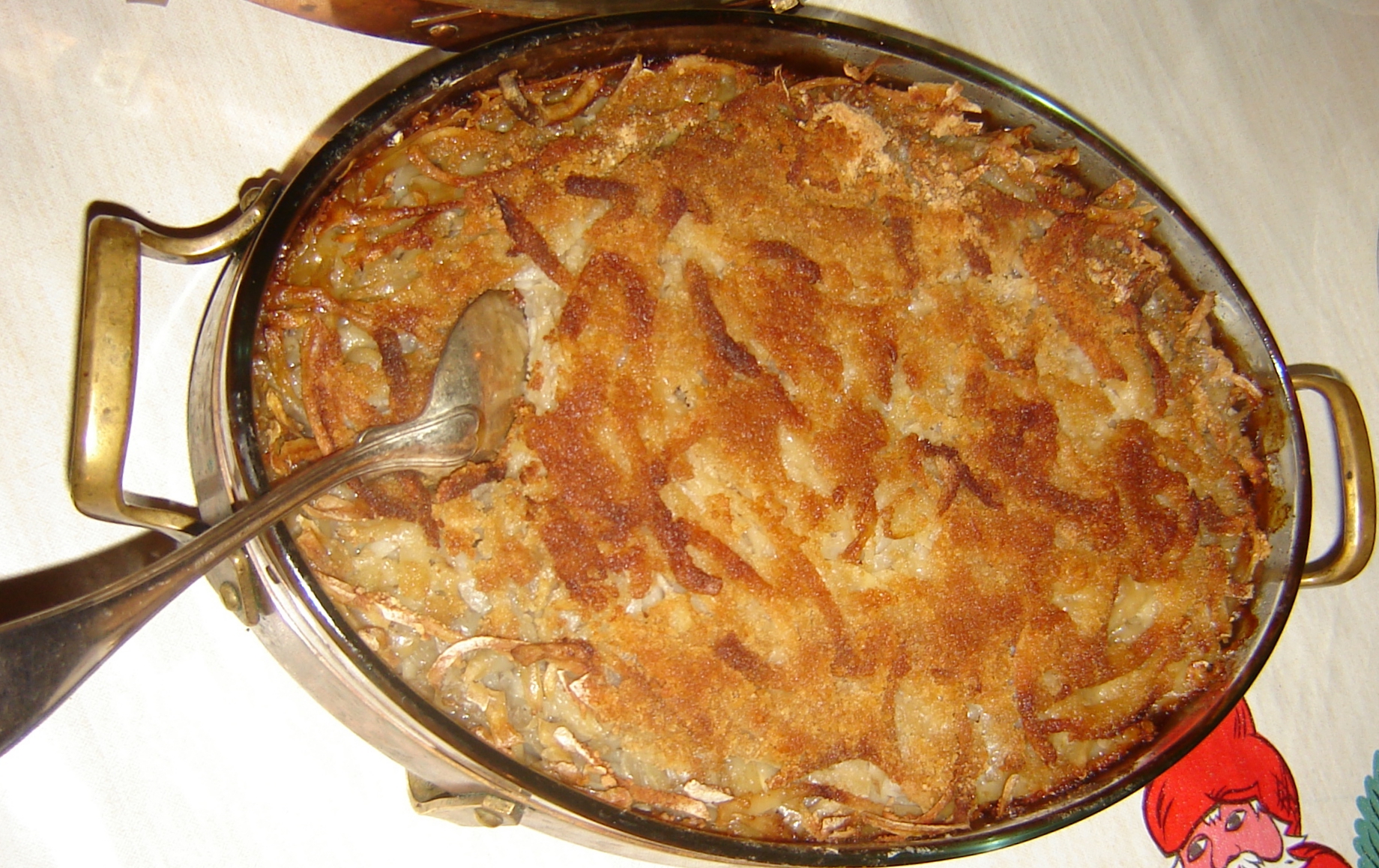 Janssons frestelse in an oval metal gratin dish with handles, the top browned and crisp, with a serving spoon resting in one corner
