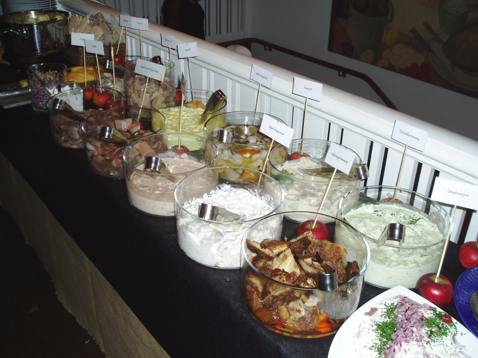A long julbord table lined with glass bowls of pickled herring in different sauces, each labelled with a small card, with red apples and a bowl of boiled potatoes