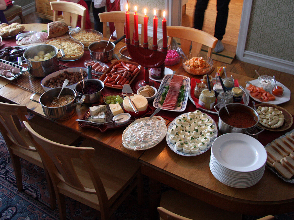A home julbord laid across a wooden dining table: platters of halved eggs, cold cuts, salmon and bread, pots of warm dishes, a stack of white plates and a row of lit red Advent candles