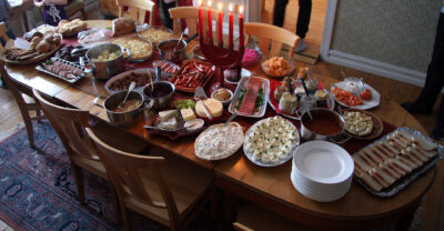 A home julbord laid across a wooden dining table: platters of halved eggs, cold cuts, salmon and bread, pots of warm dishes, a stack of white plates and a row of lit red Advent candles