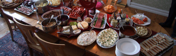 A home julbord laid across a wooden dining table: platters of halved eggs, cold cuts, salmon and bread, pots of warm dishes, a stack of white plates and a row of lit red Advent candles