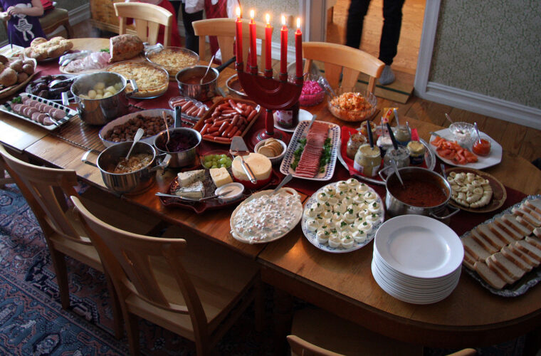 A home julbord laid across a wooden dining table: platters of halved eggs, cold cuts, salmon and bread, pots of warm dishes, a stack of white plates and a row of lit red Advent candles