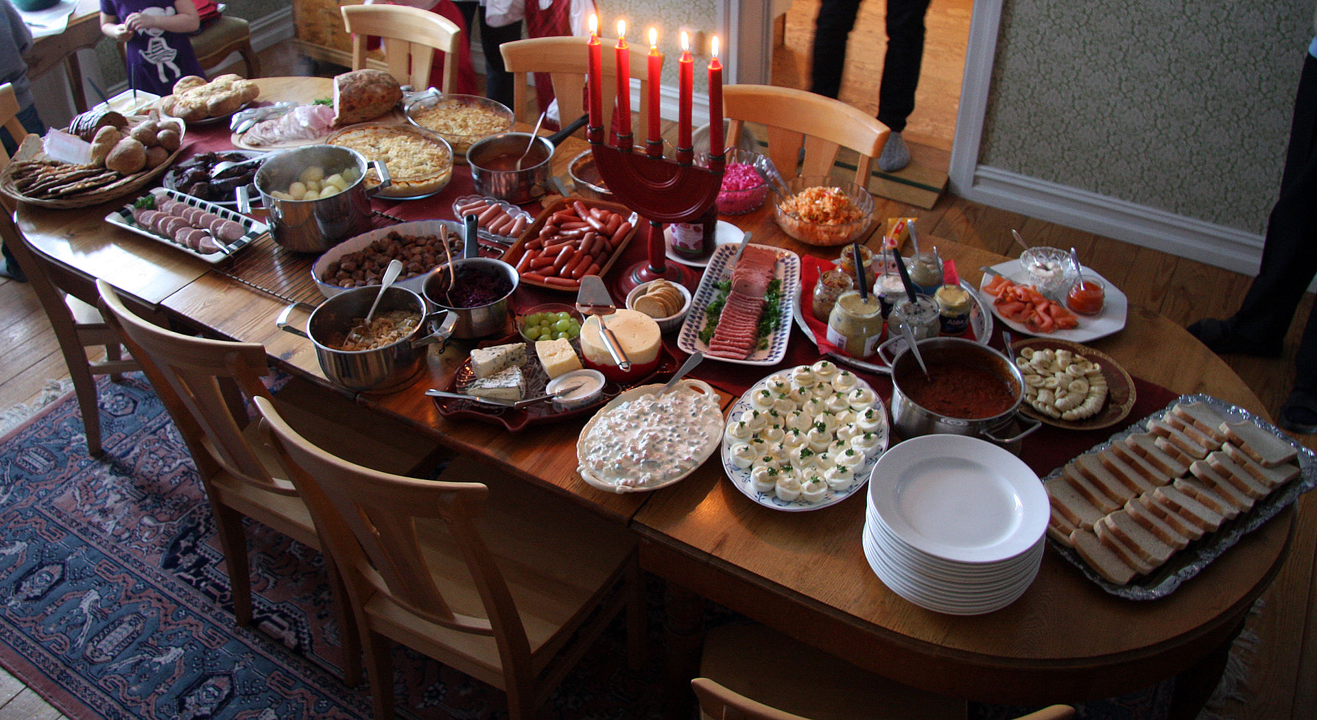 A home julbord laid across a wooden dining table: platters of halved eggs, cold cuts, salmon and bread, pots of warm dishes, a stack of white plates and a row of lit red Advent candles