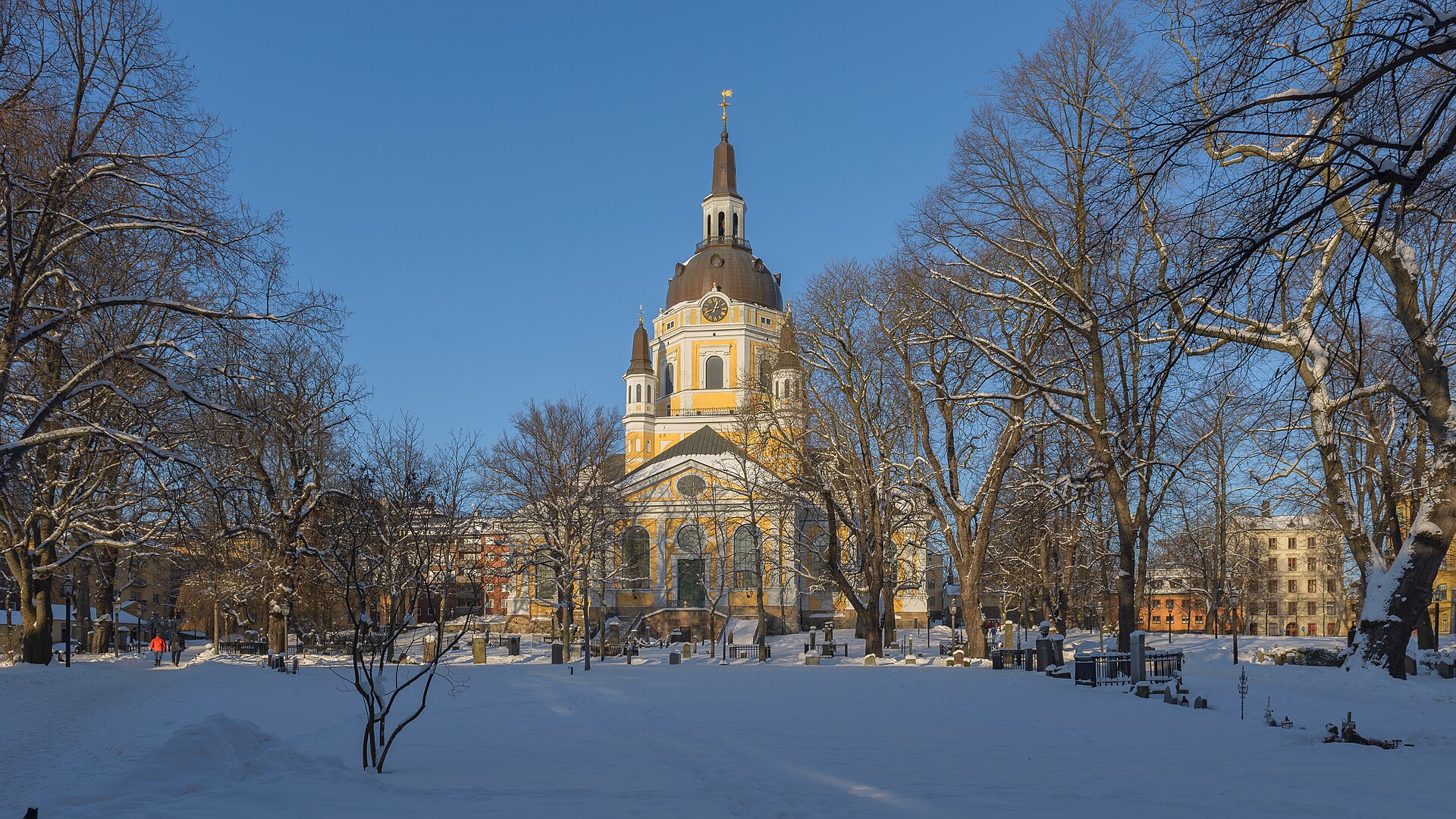The yellow Katarina Church with its dark dome and clock tower behind snow-laden trees and a snow-covered churchyard under a clear blue sky on Södermalm