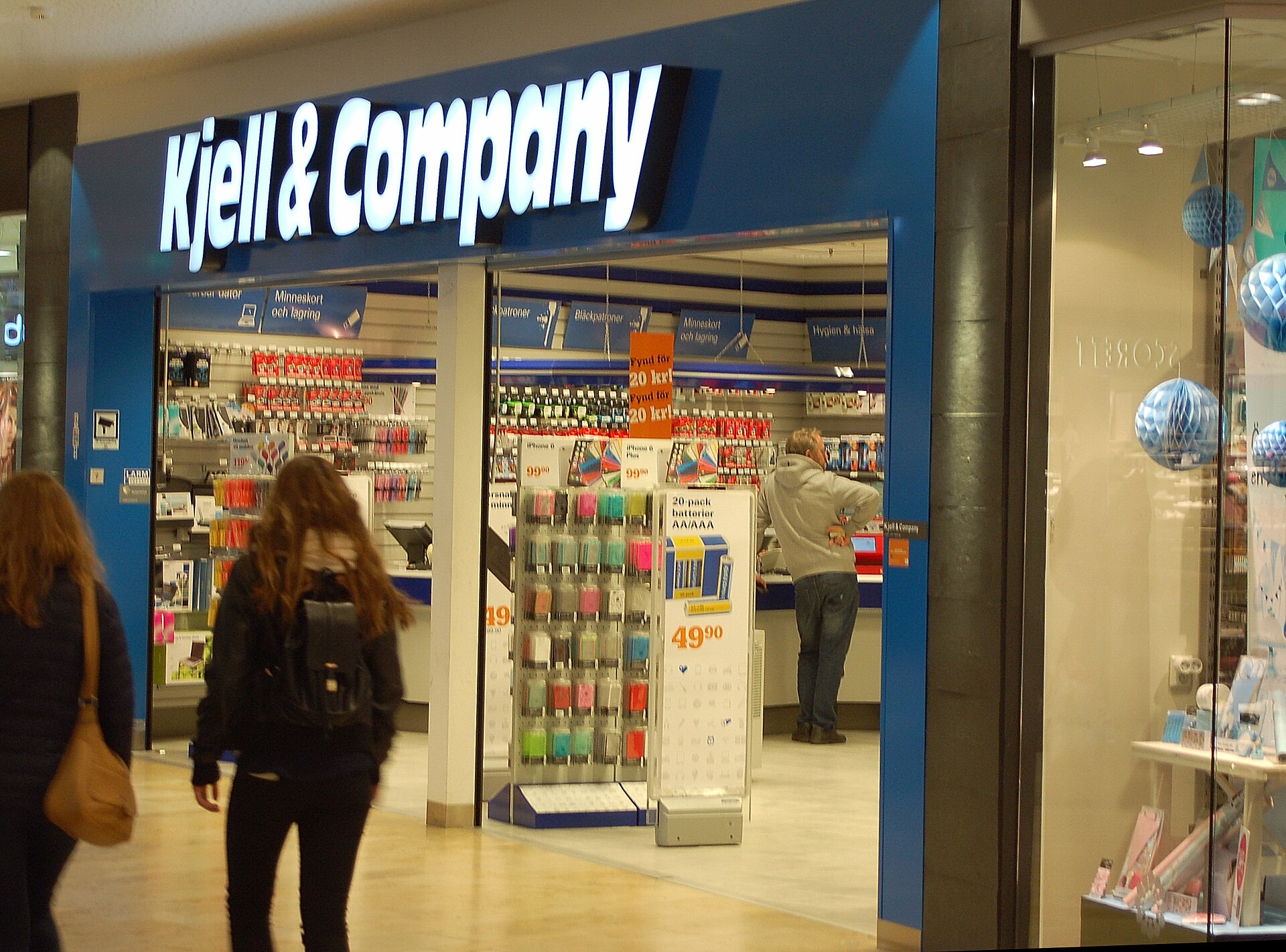 The blue-and-white shopfront of a Kjell & Company electronics store in a shopping centre, with shoppers walking past
