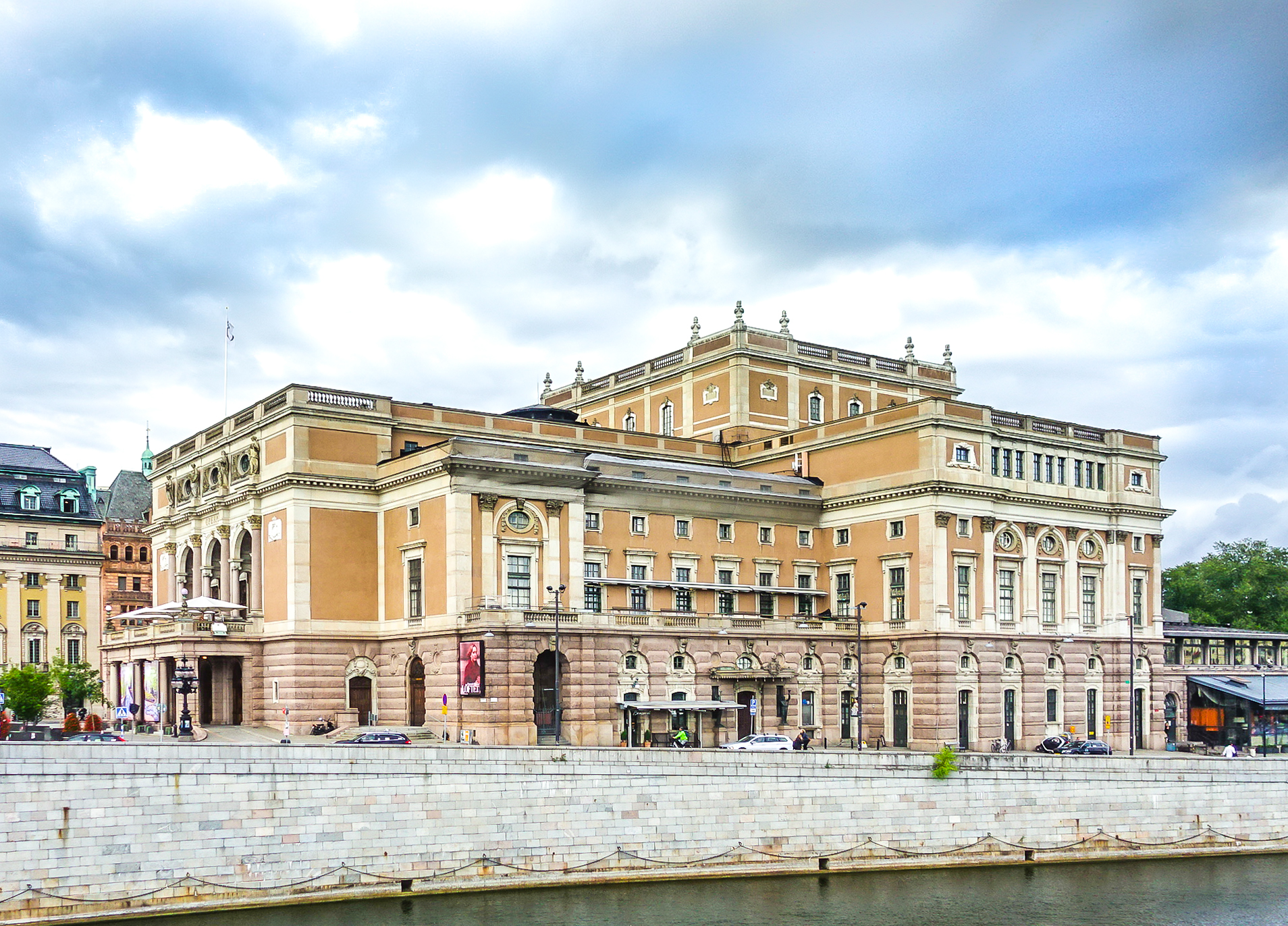 The Royal Swedish Opera house in Stockholm, a long ochre and pink stone building with arched windows, seen across the water under a cloudy sky