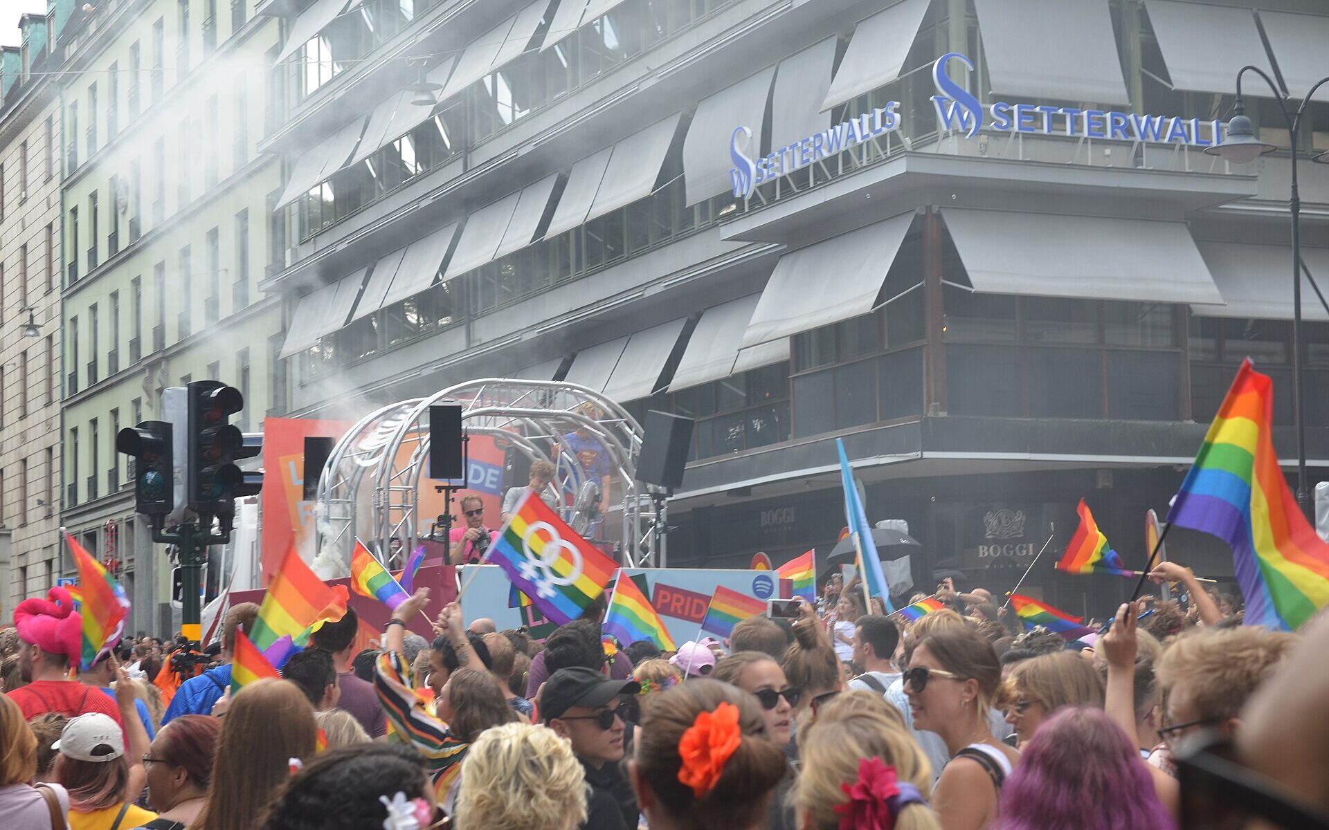 A dense crowd waving rainbow flags around a parade float at a street corner in central Stockholm during EuroPride