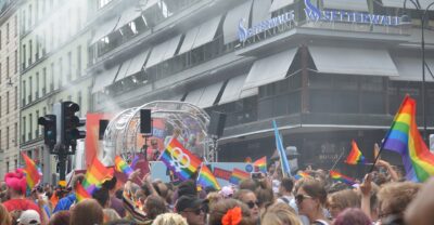 A dense crowd waving rainbow flags around a parade float at a street corner in central Stockholm during EuroPride