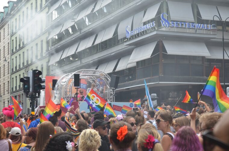 A dense crowd waving rainbow flags around a parade float at a street corner in central Stockholm during EuroPride