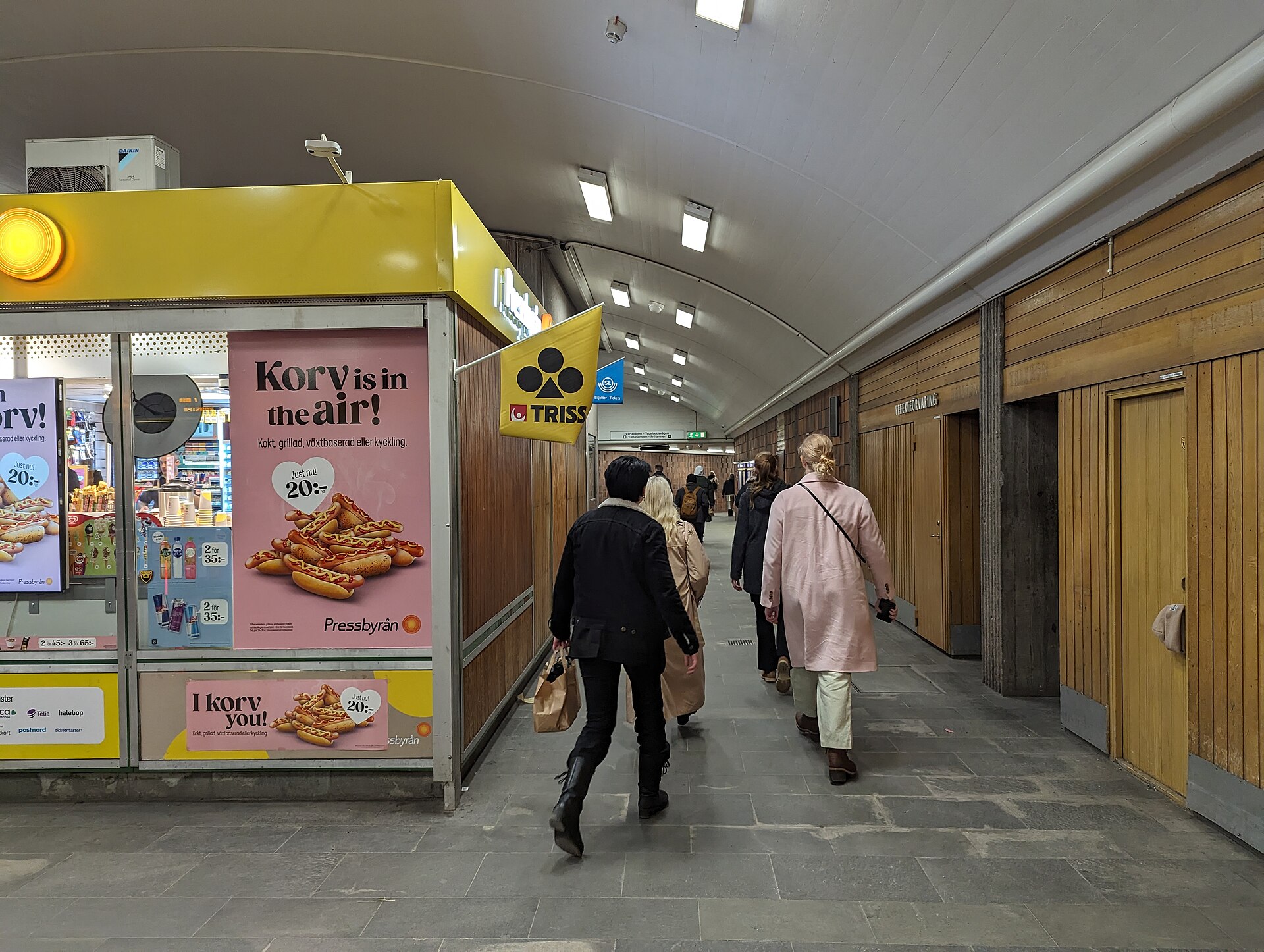 A Pressbyrån kiosk with a yellow sign and hot-dog adverts in the ticket hall of Gärdet metro station, with passengers walking past