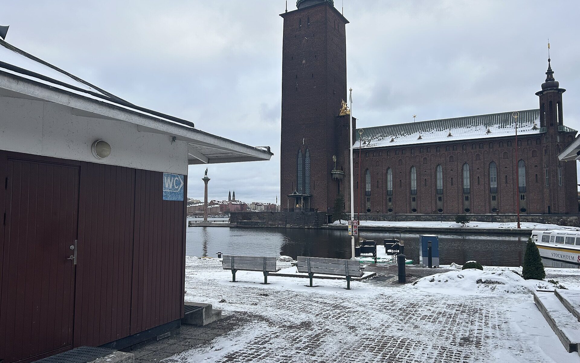 A small public toilet building on the snowy quay at Klara Mälarstrand, with Stockholm City Hall's tower across the water