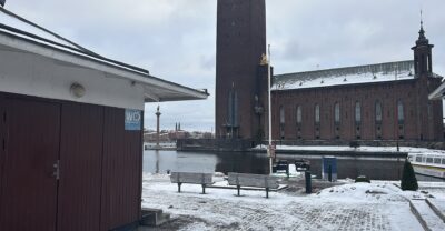 A small public toilet building on the snowy quay at Klara Mälarstrand, with Stockholm City Hall's tower across the water