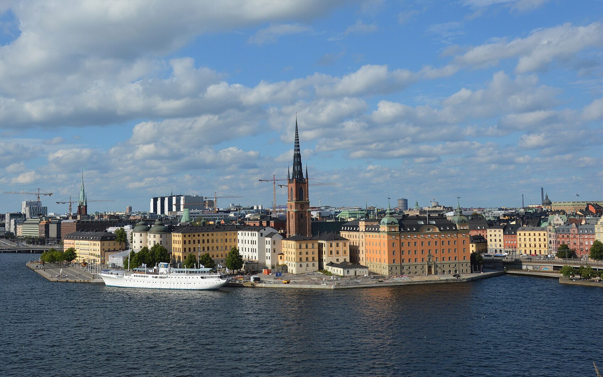 Riddarholmen's black-spired church, orange and yellow palaces and a moored white ship seen across the water from the Södermalm cliffs