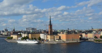 Riddarholmen's black-spired church, orange and yellow palaces and a moored white ship seen across the water from the Södermalm cliffs