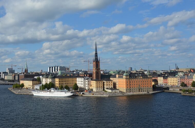 Riddarholmen's black-spired church, orange and yellow palaces and a moored white ship seen across the water from the Södermalm cliffs