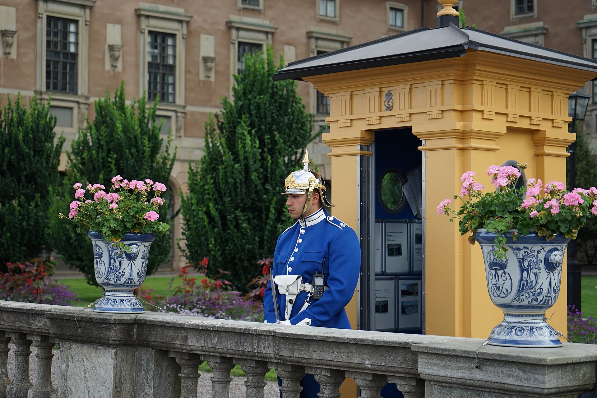 A Royal Guard sentry in a blue uniform and polished spiked helmet beside a yellow sentry box between two blue-and-white flower urns at the Royal Palace