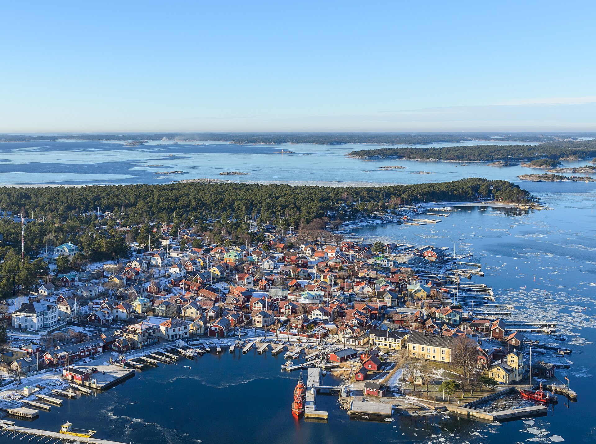 Aerial view of Sandhamn in February, with red and yellow wooden houses around a harbour, pine forest and broken ice drifting on the blue water of the archipelago