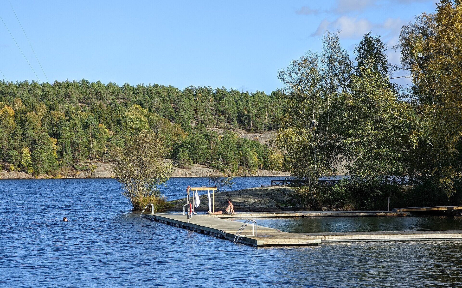A bather resting on a wooden jetty with towels hanging on a rack while another swims in the blue lake Källtorpssjön at Hellasgården, surrounded by autumn forest