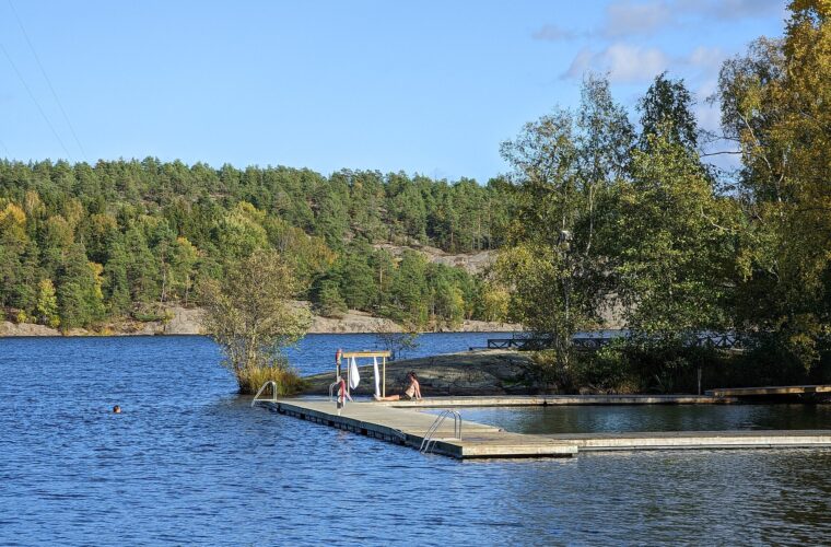 A bather resting on a wooden jetty with towels hanging on a rack while another swims in the blue lake Källtorpssjön at Hellasgården, surrounded by autumn forest
