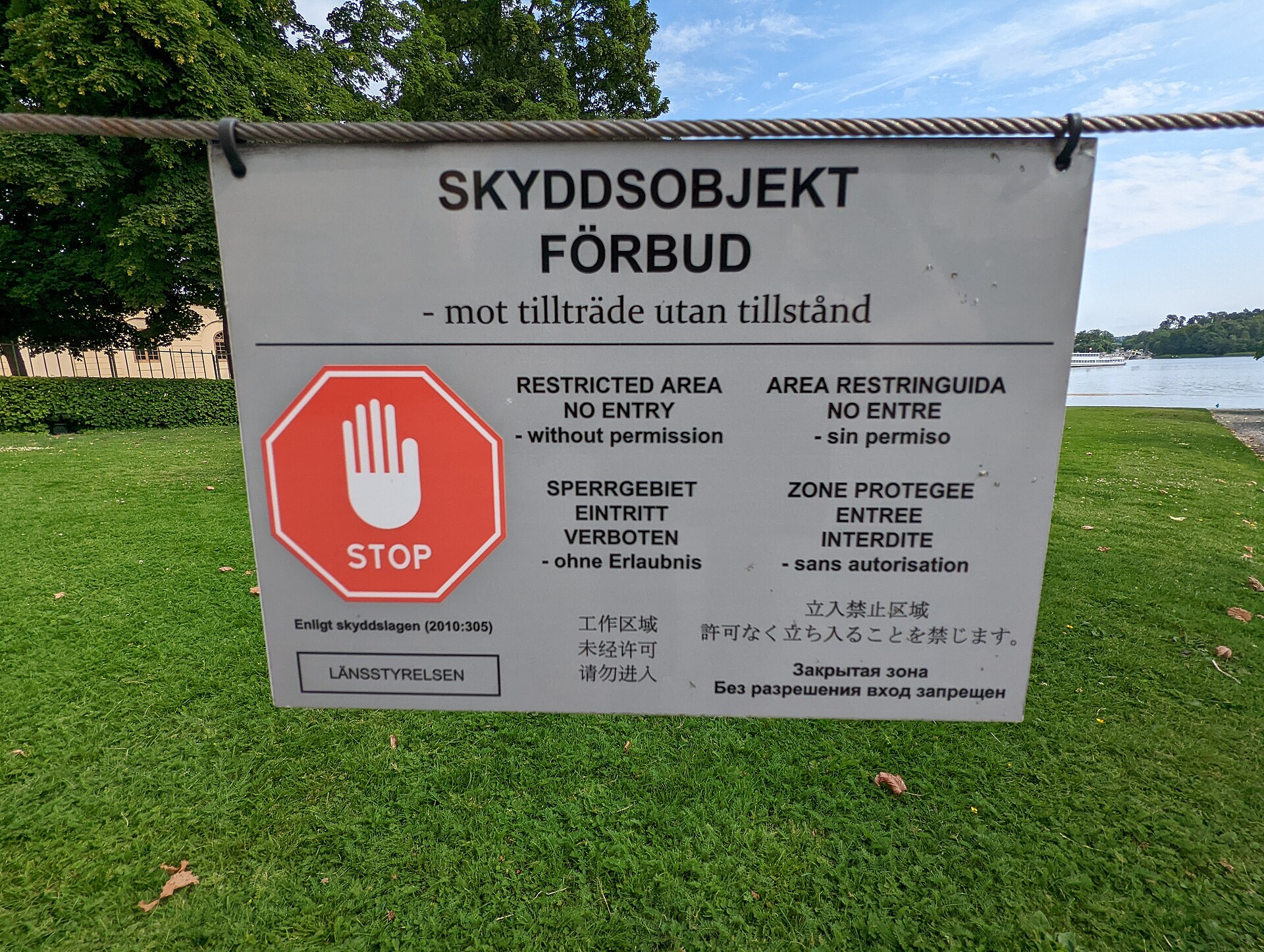 A multilingual skyddsobjekt sign hung on a wire at Drottningholm Palace, with a red stop hand, forbidding entry without permission under the Protection Act 2010:305