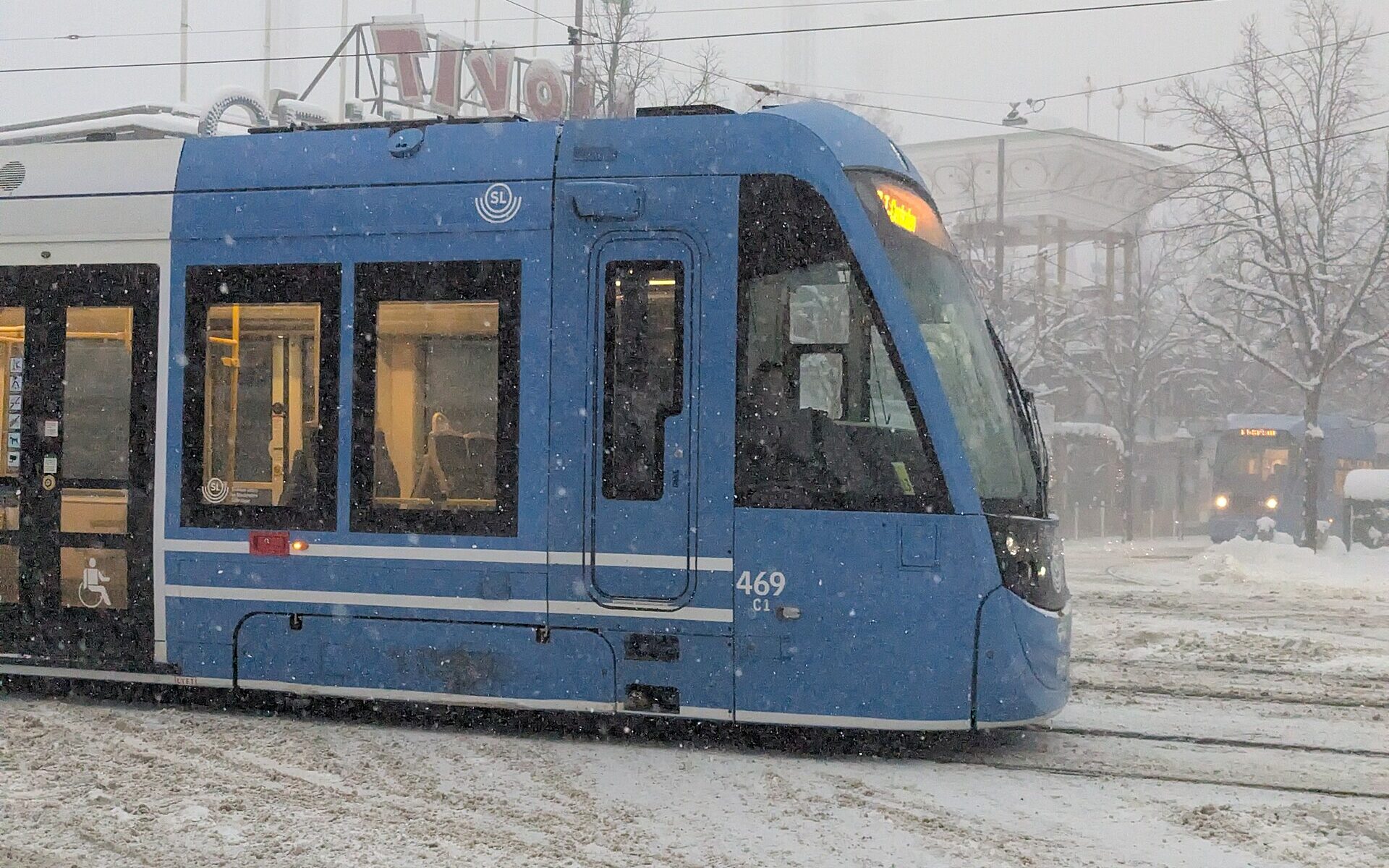 A blue low-floor SL tram with a wheelchair symbol on its door passing Gröna Lund on Djurgården in falling snow
