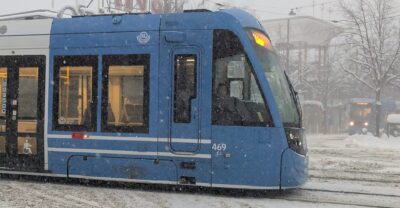 A blue low-floor SL tram with a wheelchair symbol on its door passing Gröna Lund on Djurgården in falling snow