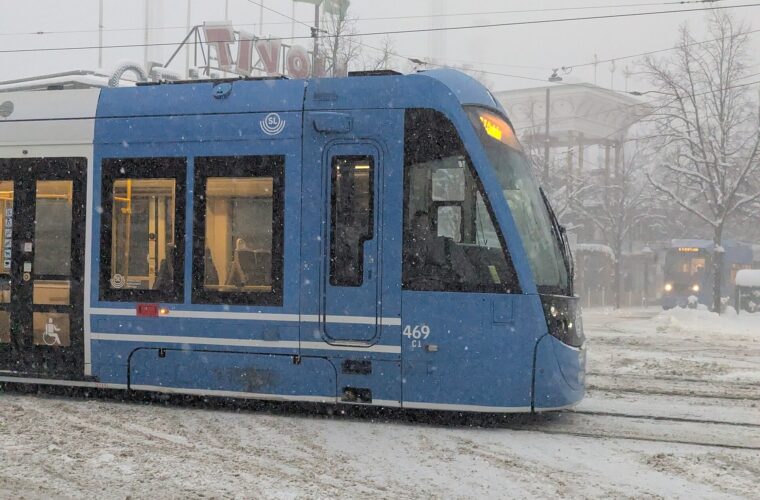 A blue low-floor SL tram with a wheelchair symbol on its door passing Gröna Lund on Djurgården in falling snow