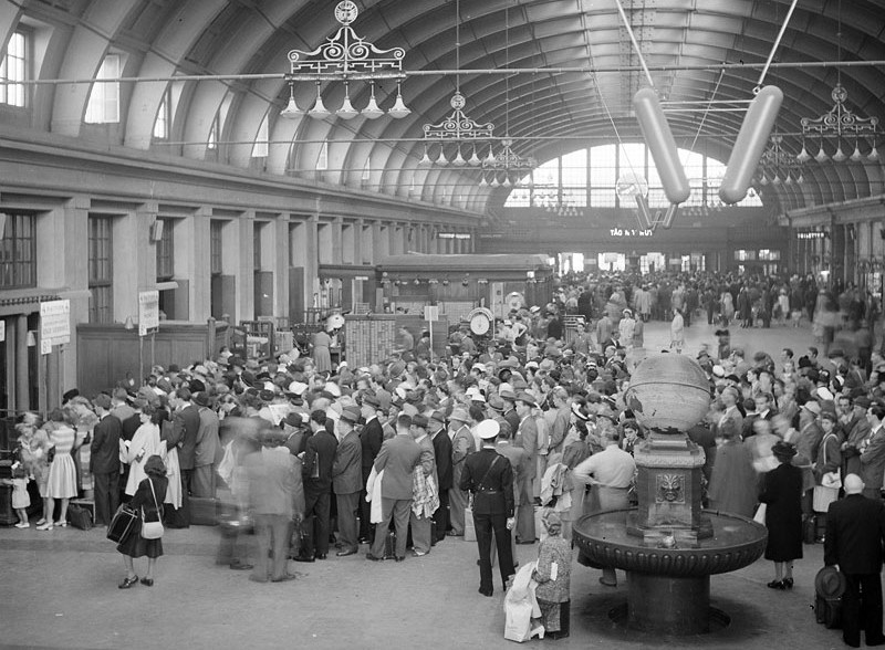 Black-and-white photograph of a crowded Stockholm Central station hall in the 1940s, with travellers in hats and coats under a vaulted ceiling hung with chandeliers