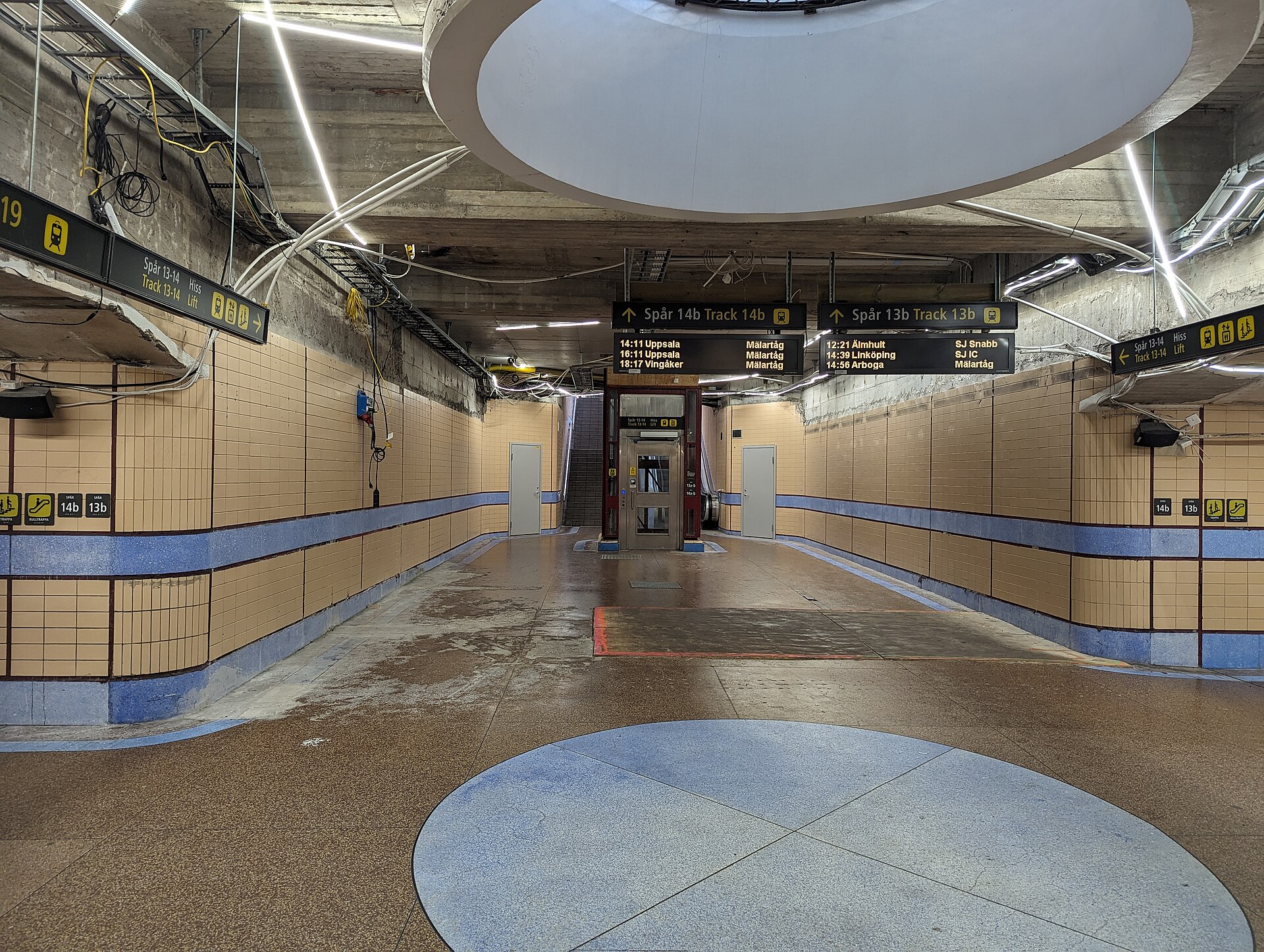 An underground passage at Stockholm Central with a glass lift to tracks 13 and 14, bilingual Hiss and Lift signs and departure boards overhead