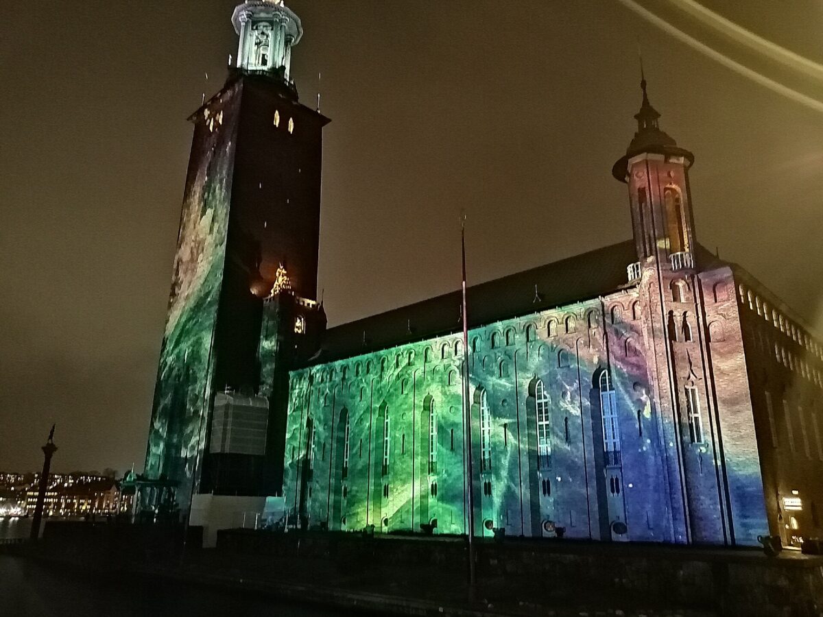 Stockholm City Hall at night, its brick tower and long facade washed in green, blue and violet projected light during Nobel Week Lights