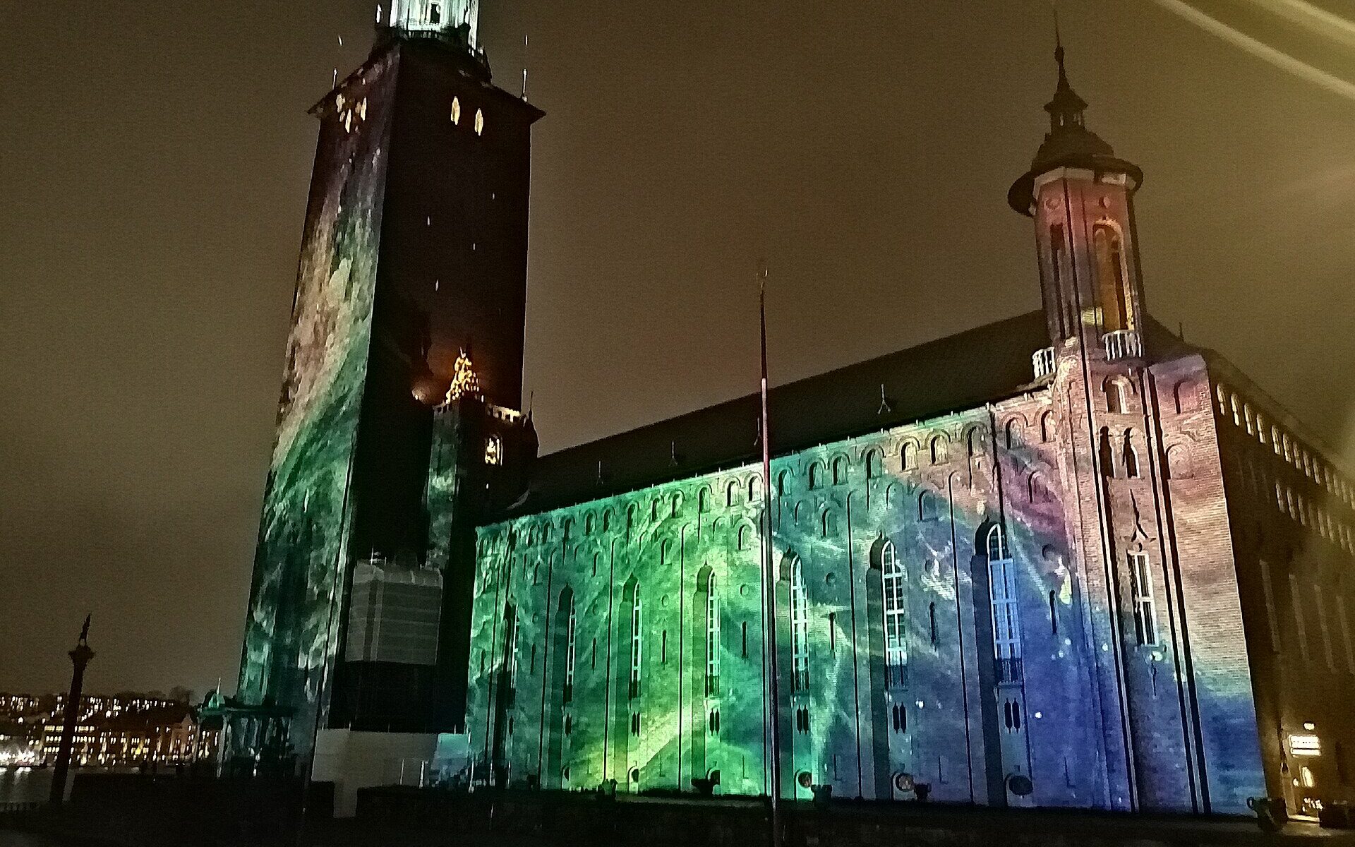 Stockholm City Hall at night, its brick tower and long facade washed in green, blue and violet projected light during Nobel Week Lights