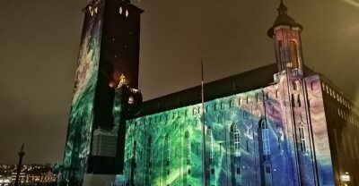 Stockholm City Hall at night, its brick tower and long facade washed in green, blue and violet projected light during Nobel Week Lights